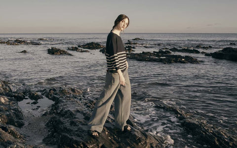 Les Racines du Ciel Baby-Alpaka Pullover mit Streifenmuster – Frau auf Felsen am Meer, dunkler Strickpullover mit weißen Streifen und weiter Hose